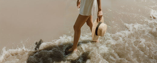 Woman walking along the beach holding a sunhat, showcasing essential travel skincare tips for protecting skin from sun exposure.