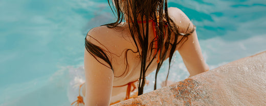 A woman with wet hair sitting on the edge of a pool, partially submerged in water, enjoying a sunny day by the poolside.